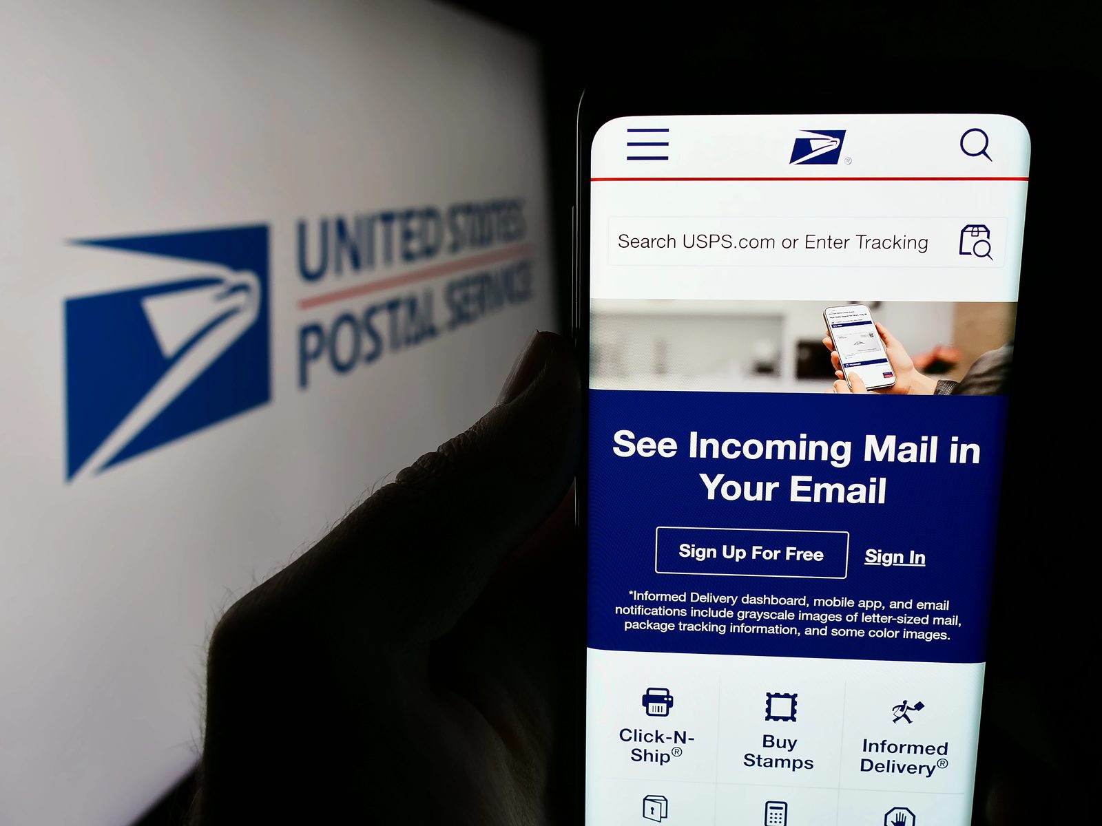 Hand holding a phone showing the USPS website with Informed Delivery sign-up, in front of a USPS logo